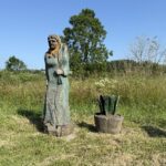 Holzskulptur einer Bäuerin auf weiter Sommerwiese mit Feldern im Hintergrund