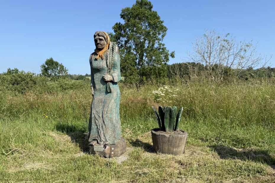 Holzskulptur einer Bäuerin auf weiter Sommerwiese mit Feldern im Hintergrund