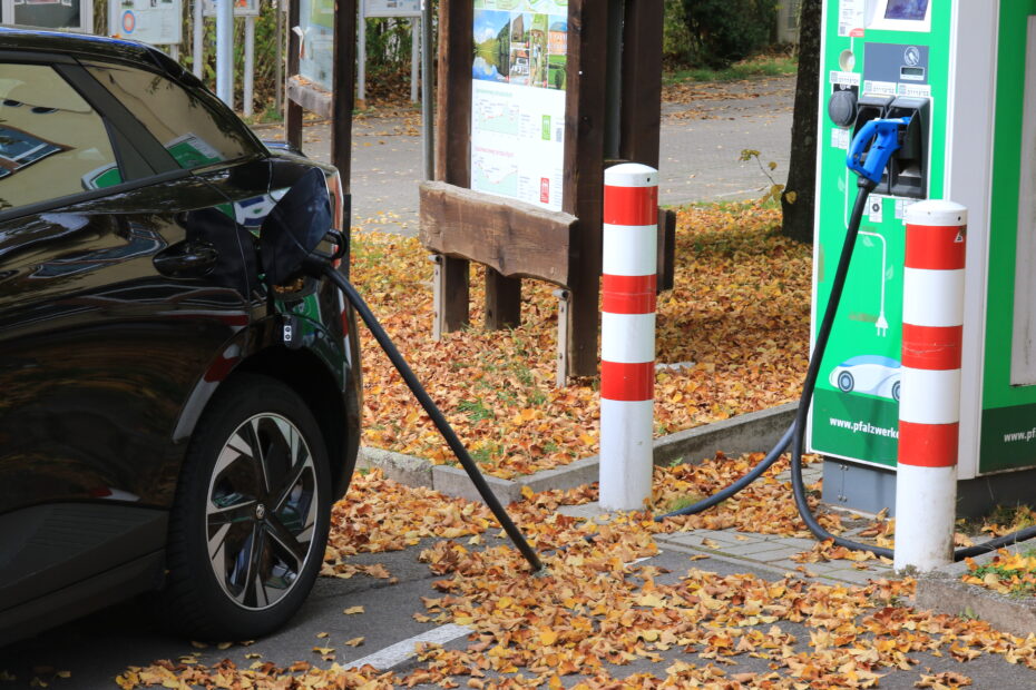 E-Auto lädt an der Ladesäule in Trippstadt; Herbstlaub bedeckt den Boden, Infotafeln im Hintergrund – Startpunkt nahe der Karlstalschlucht.