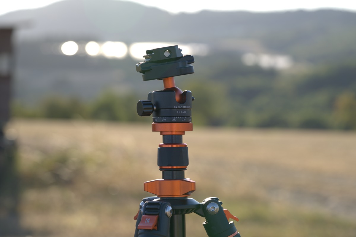 K&F Concept Kugelkopf-Stativ in Orange-Schwarz im Vordergrund, unscharfes Hügelpanorama im Hintergrund