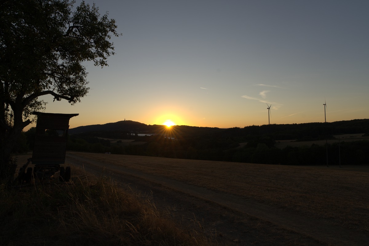 Sonnenuntergang hinter Hügel mit Denkmal, Windräder rechts, Feldweg und Hochsitz als Silhouette – Sonnenfinsternis Wandertour